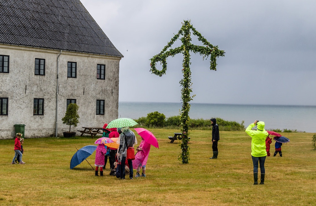 Midsommarfirande utomhus med en traditionell midsommarstång och människor med paraplyer i regnigt väder.